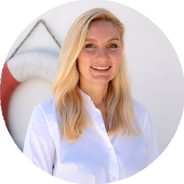 Porträtfoto von Anne Katrin Fischer, Executive Partner von Emocean Cruises. Eine lächelnde Frau mit schulterlangem, blonden Haar. Sie trägt eine weiße Bluse und steht vor einem weißen Hintergrund mit einem Rettungsring, der teilweise sichtbar ist.